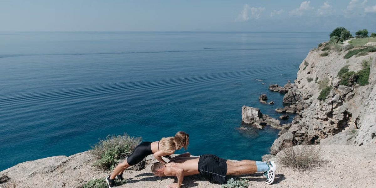 A couple performs a partner workout on a scenic cliffside, overlooking the ocean.